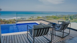 A rooftop infinity pool with two lounge chairs and a table, overlooking a beach and ocean.