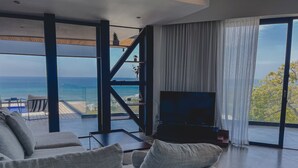 A modern living room with a large TV, grey sofas, and a view of the ocean.