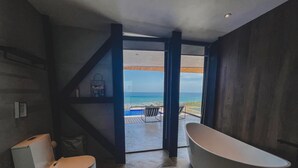A modern bathroom with a freestanding bathtub, a toilet, and a view of the ocean through an open doorway.