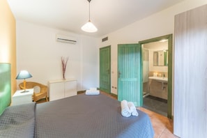 Un dormitorio con cama, mesita de noche, lámpara, silla, puerta y un baño visible a través de una puerta abierta.