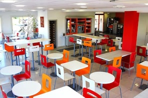 A cafeteria with multiple tables and chairs, a counter with a beverage dispenser, and a refrigerator.