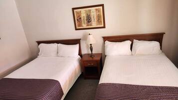 A hotel room with two single beds, each with a wooden headboard, white linens, and a dark striped bedspread. There is a nightstand with a lamp between the beds, and a framed botanical print on the wall above the nightstand.
