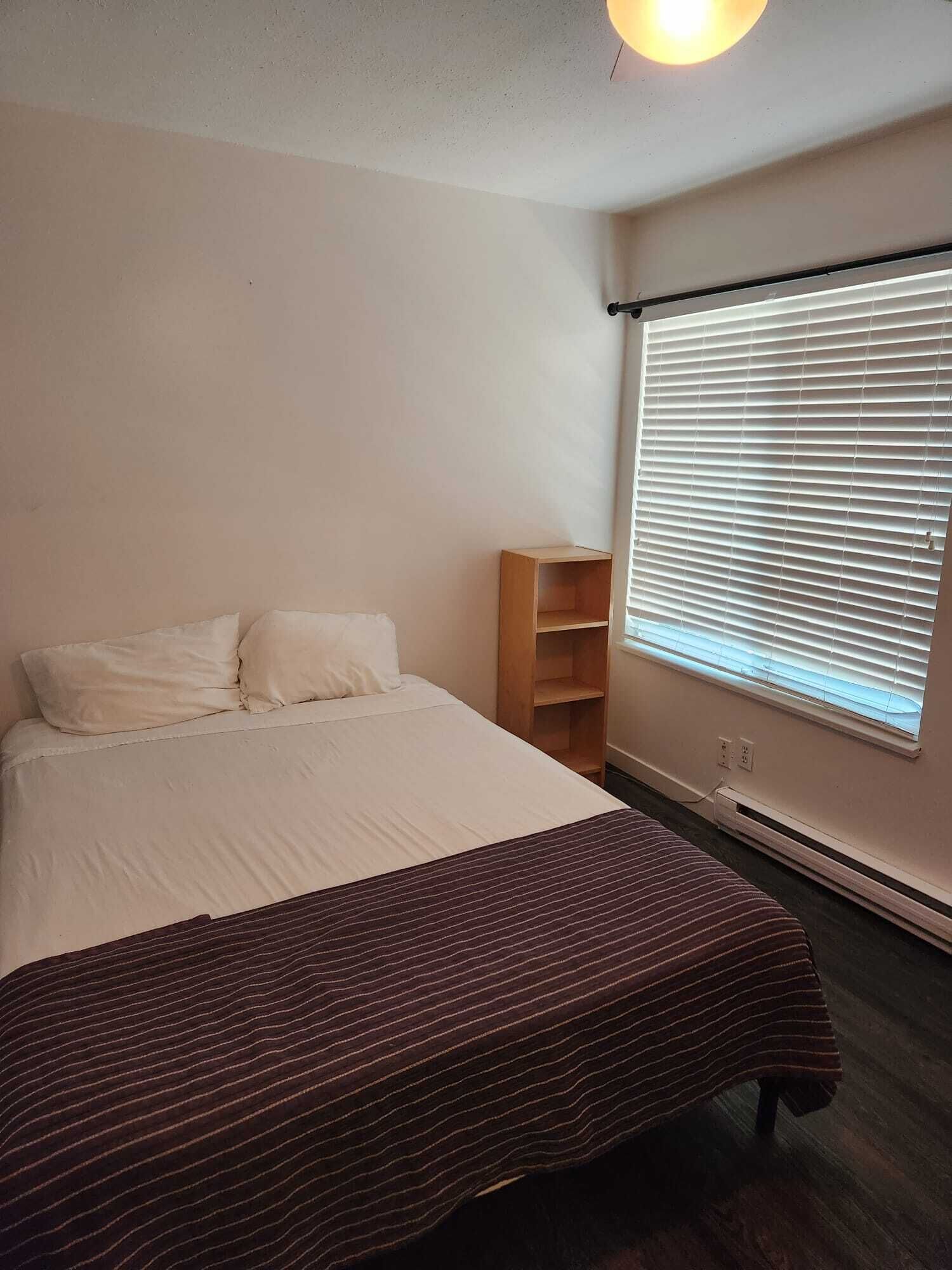 Un dormitorio con cama, una mesita de noche de madera, una ventana con persianas y un calentador fijado a la pared.