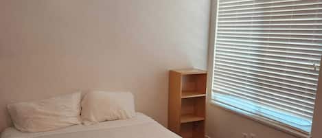 Un dormitorio con cama, una mesita de noche de madera, una ventana con persianas y un calentador fijado a la pared.