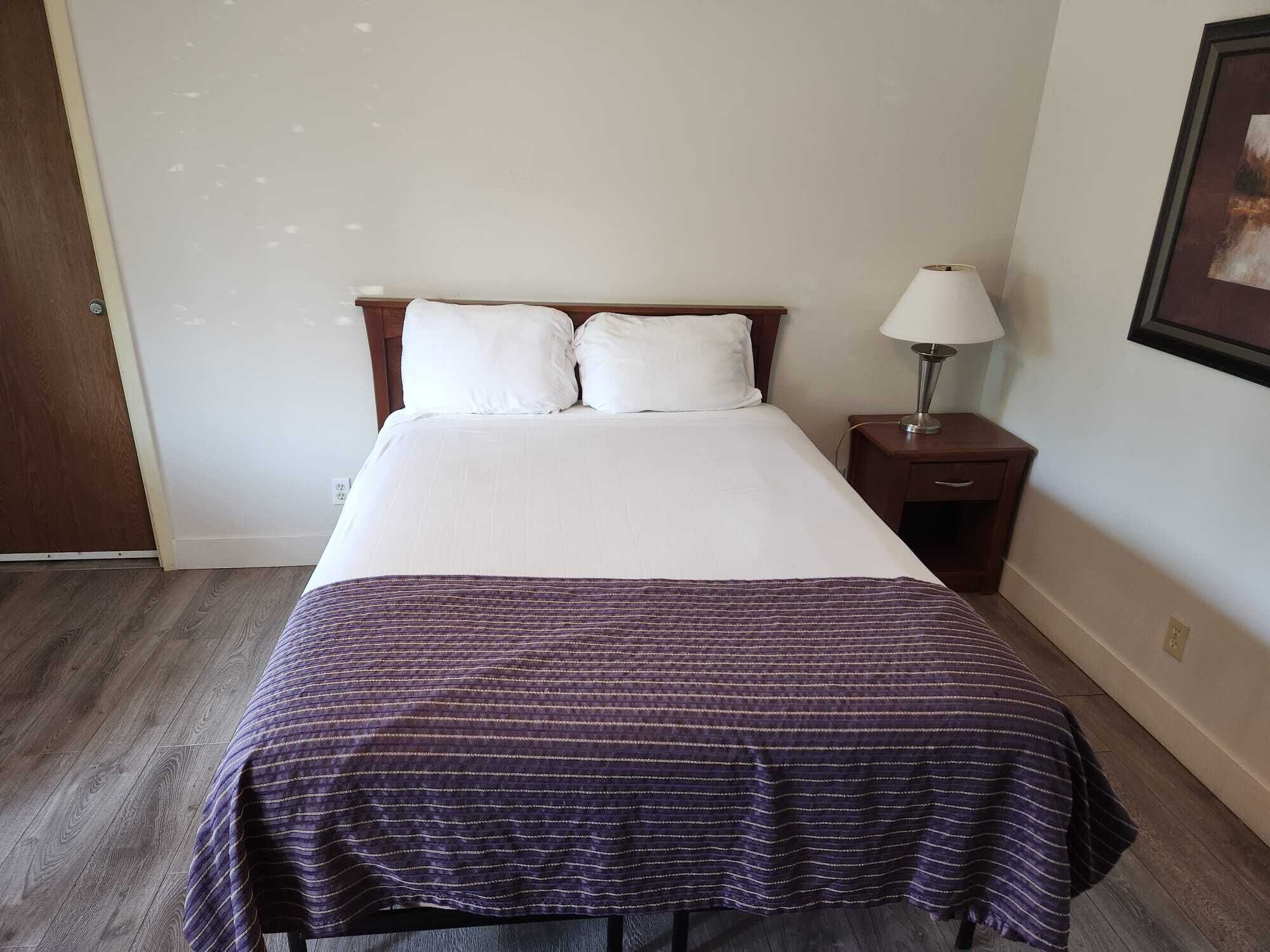Ein ordentlich bezogenes Bett mit weißer Daunendecke und einer violett gestreiften Tagesdecke, ein Nachttisch aus Holz mit einer Lampe und ein gerahmtes Bild an der Wand.