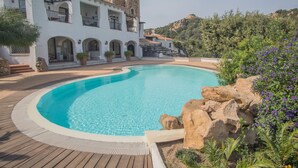 A swimming pool with a wooden deck, a white building with arches and balconies, and a mountainous backdrop. - Beautiful Il Giardino Degli Oleandri 1 Bedroom Apartment B2 Sleeps 2 Persons (Arzachena)