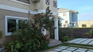 A two-story house with a balcony, a garden with a paved walkway, and a water tank in the background. - Beach Rustic Condo (Nassau)