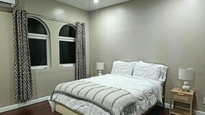 A bedroom with a bed, two bedside tables, a window with curtains, and a ceiling fan. - Beach Rustic Condo (Nassau)
