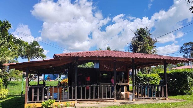A rustic wooden structure with a red roof, surrounded by lush greenery and a clear blue sky.