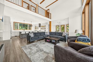 A spacious living area with a high ceiling, a kitchen with bar seating, and large windows offering views of greenery. - Fernie Alpine Chalet (Fernie)