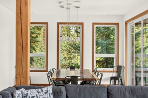 A dining area with a wooden table and chairs, large windows with a view of trees, and a hanging light fixture.