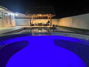 Une piscine éclairée de lumières bleues, une structure à toit de chaume et un espace détente avec des coussins.