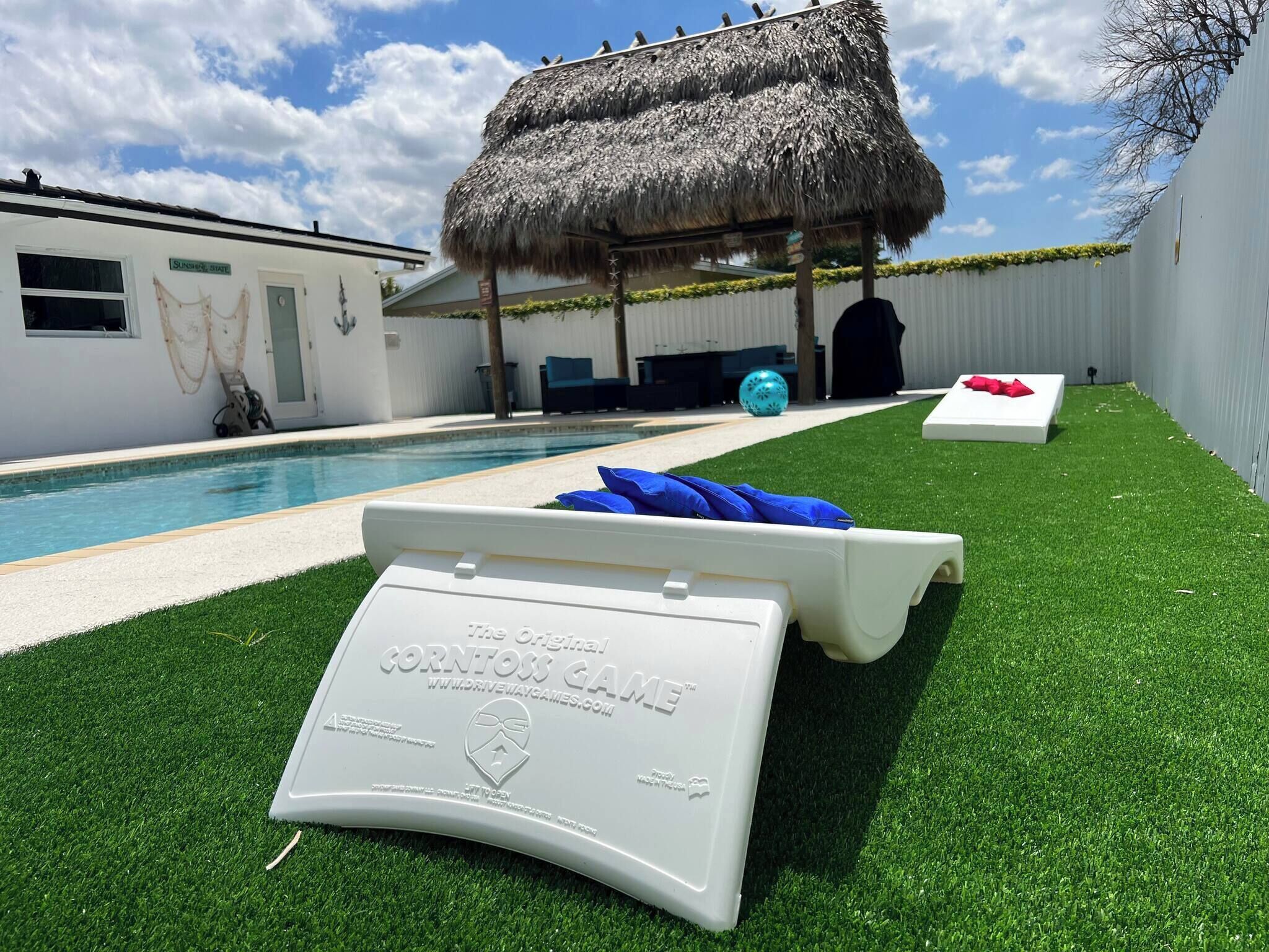 A pool area with a thatched roof, artificial grass, lounge chairs, and a sign for 'The Original CornToss Game.'