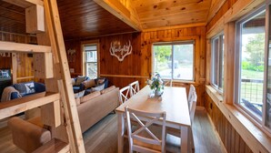 L’intérieur d’un chalet en bois comprend une table à manger, des chaises blanches, un canapé avec des coussins et une grande fenêtre donnant sur l’extérieur.