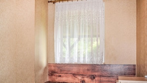 A room with a wooden headboard, a window with sheer white curtains, and a view of greenery outside.