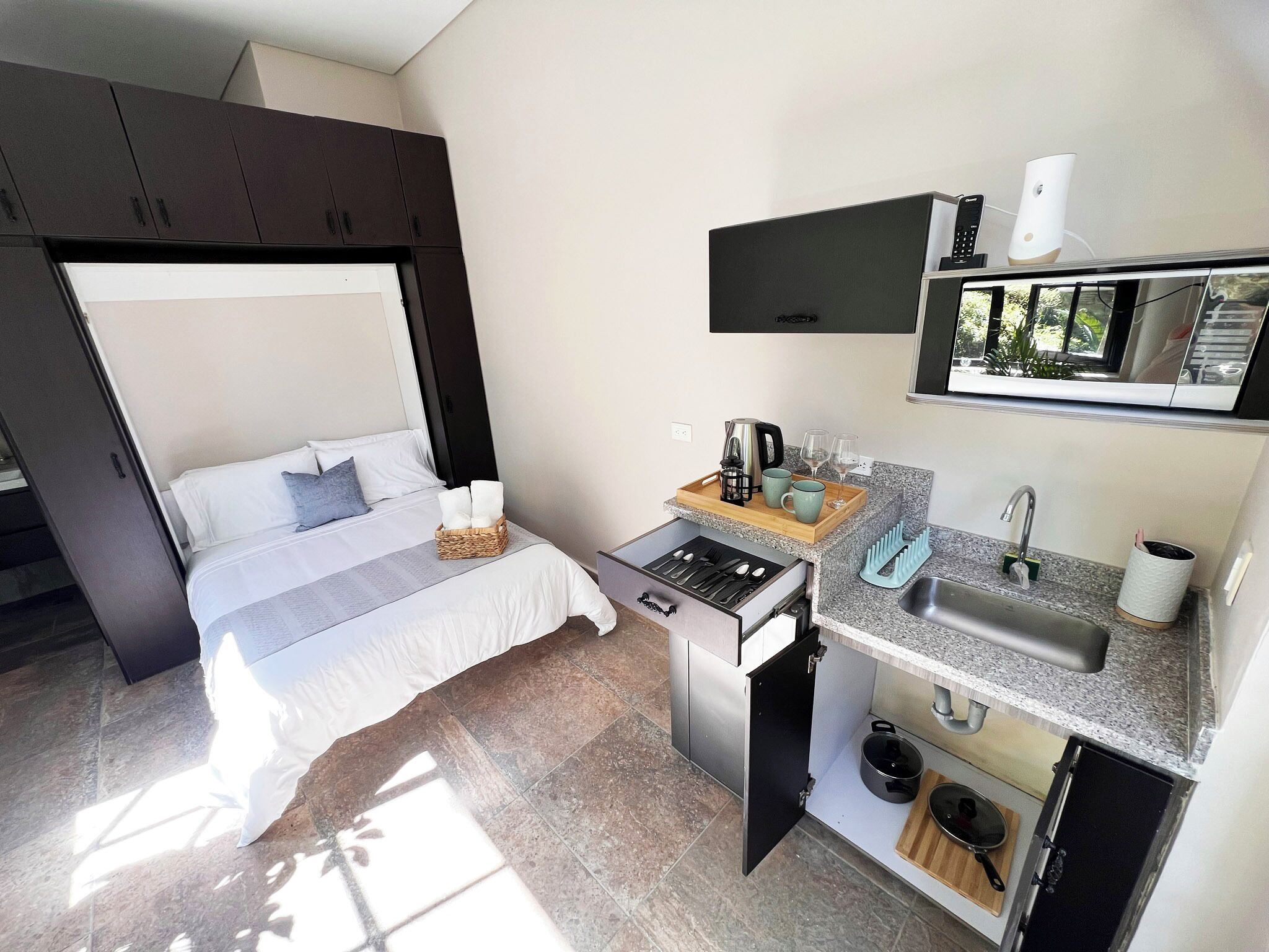 Habitación de hotel pequeña con cama, una kitchenette y un televisor montado en la pared.