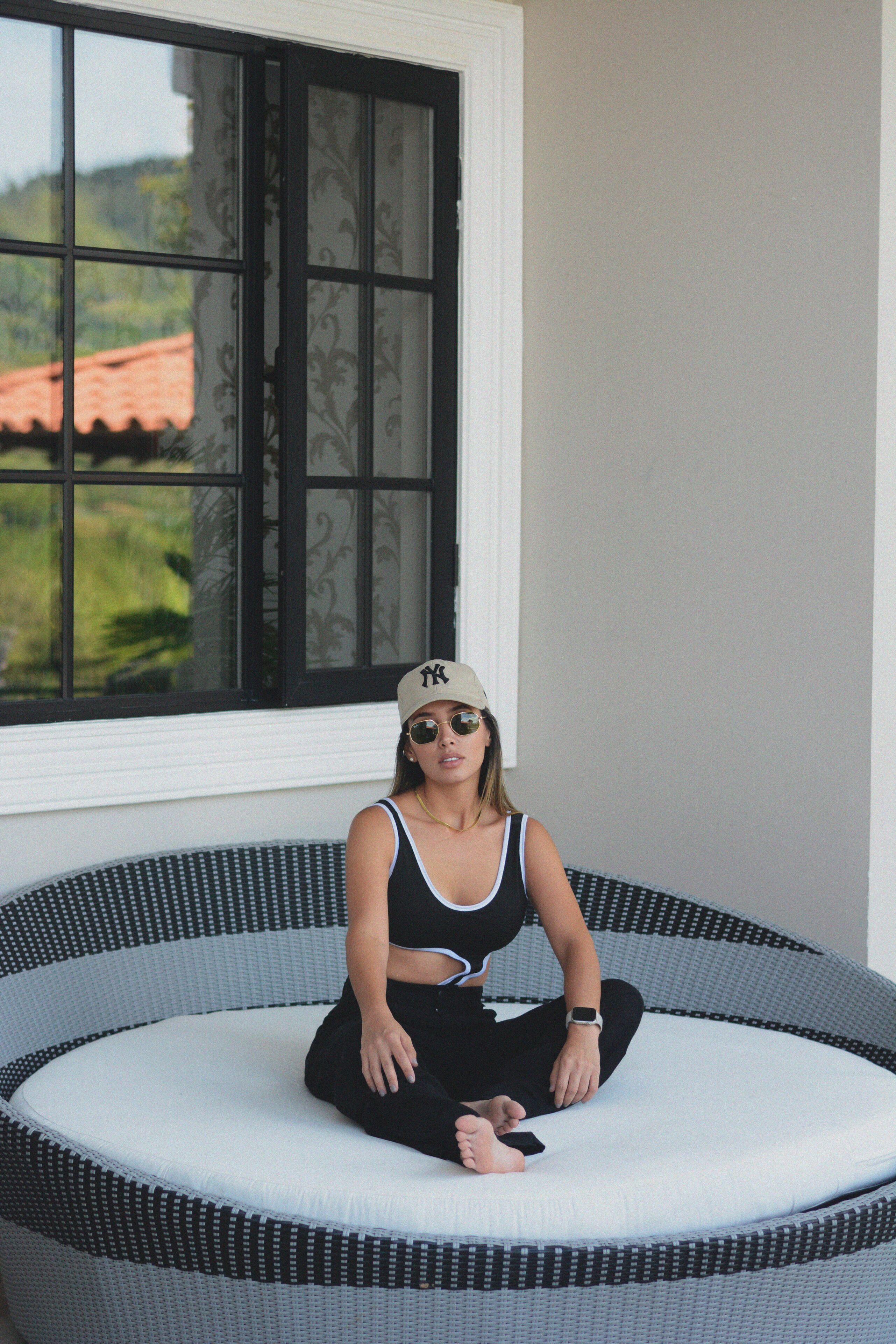 Una persona sentada en una silla circular de mimbre con un cojín blanco, vestida con una camiseta de tirantes negra, pantalones negros y una gorra color beige.