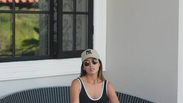 Una persona sentada en una silla circular de mimbre con un cojín blanco, vestida con una camiseta de tirantes negra, pantalones negros y una gorra color beige.