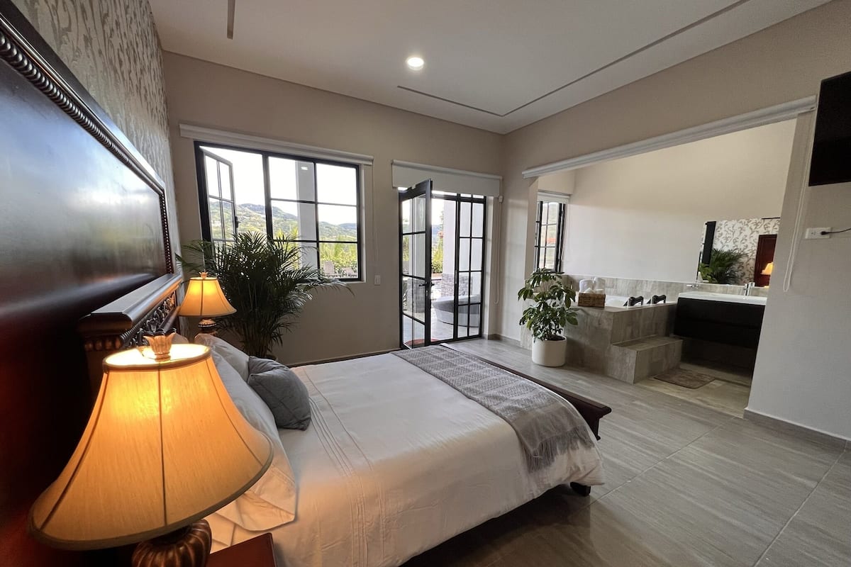 A bedroom with a bed, bedside lamps, a potted plant, and a view of the outdoors through large windows.
