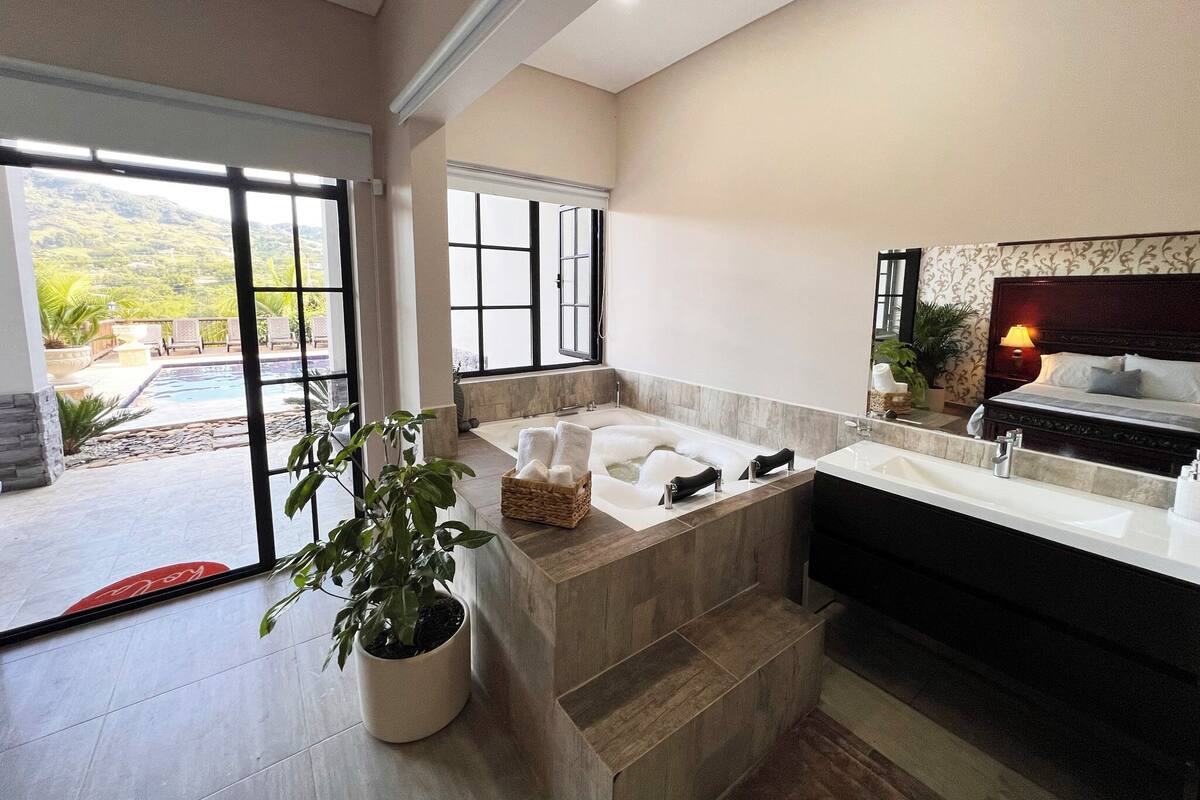 A modern bathroom with a large bathtub, a separate shower area, and a sink. There is a potted plant in the corner and a view of a pool and mountains through the windows.