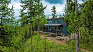 A cabin surrounded by dense forest, with a deck and outdoor seating area. - One of a kind views of Glacier National Park! Cascade Cabin at Thunderbird Ridge. (Polebridge, Montana)