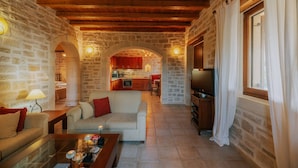 A cozy living room with a sofa, coffee table, and TV, connected to a kitchen with a stone wall and wooden ceiling. - Villa Arianna With Breathtaking View Near Rethymno (Rethymno)