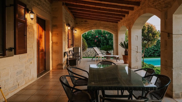 A covered outdoor patio with a table and chairs, stone walls, wooden beams, and a view of a pool. - Villa Arianna With Breathtaking View Near Rethymno (Rethymno)