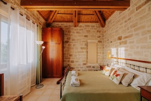 Une chambre avec un mur en pierre, des poutres en bois au plafond, un lit avec des oreillers, une table de chevet et une commode.