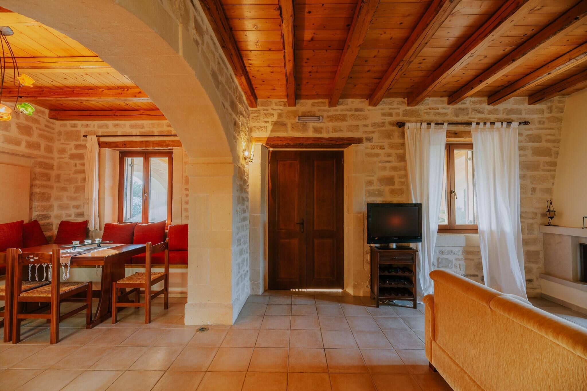 A cozy room with a stone wall, wooden ceiling beams, a dining table with chairs, a sofa, a television, and a fireplace.