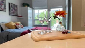 Une pièce comprenant un lit, une table en bois avec deux verres à vin et un vase avec une fleur, ainsi qu’une fenêtre donnant sur un paysage verdoyant.