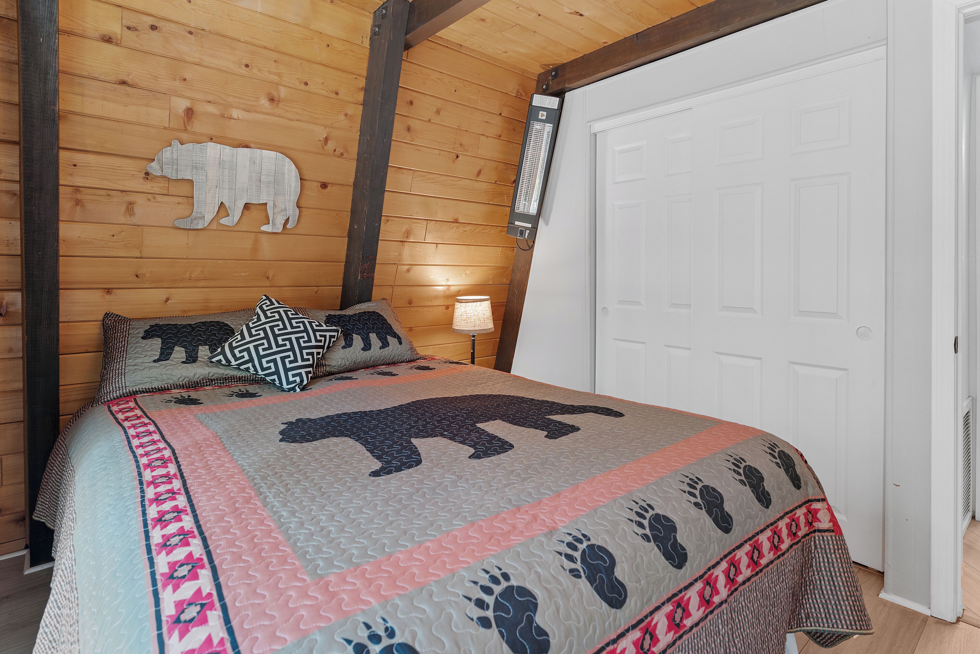 Un dormitorio de cabaña de madera con una cama que presenta un edredón con estampado de oso, una puerta blanca y una mesita de noche con una lámpara.