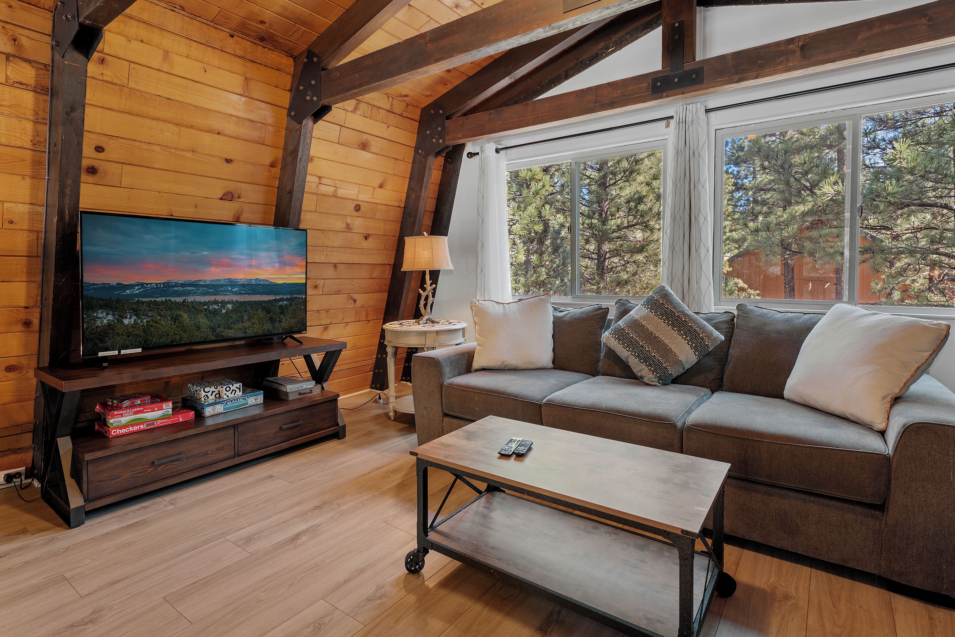 A cozy living room with a wooden ceiling, a large flat-screen TV, a sofa with cushions, a coffee table, and a window with a view of trees.