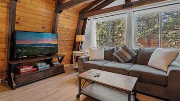 A cozy living room with a wooden ceiling, a large flat-screen TV, a sofa with cushions, a coffee table, and a window with a view of trees.