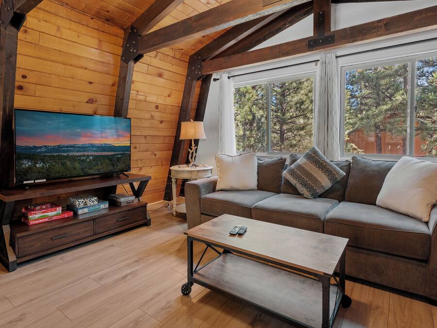 A cozy living room with a wooden ceiling, a large flat-screen TV, a sofa with cushions, a coffee table, and a window with a view of trees.