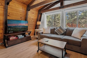 Una sala de estar acogedora con techo de madera, un televisor de pantalla plana grande, un sofá con cojines, una mesa de centro y una ventana con vista a árboles.