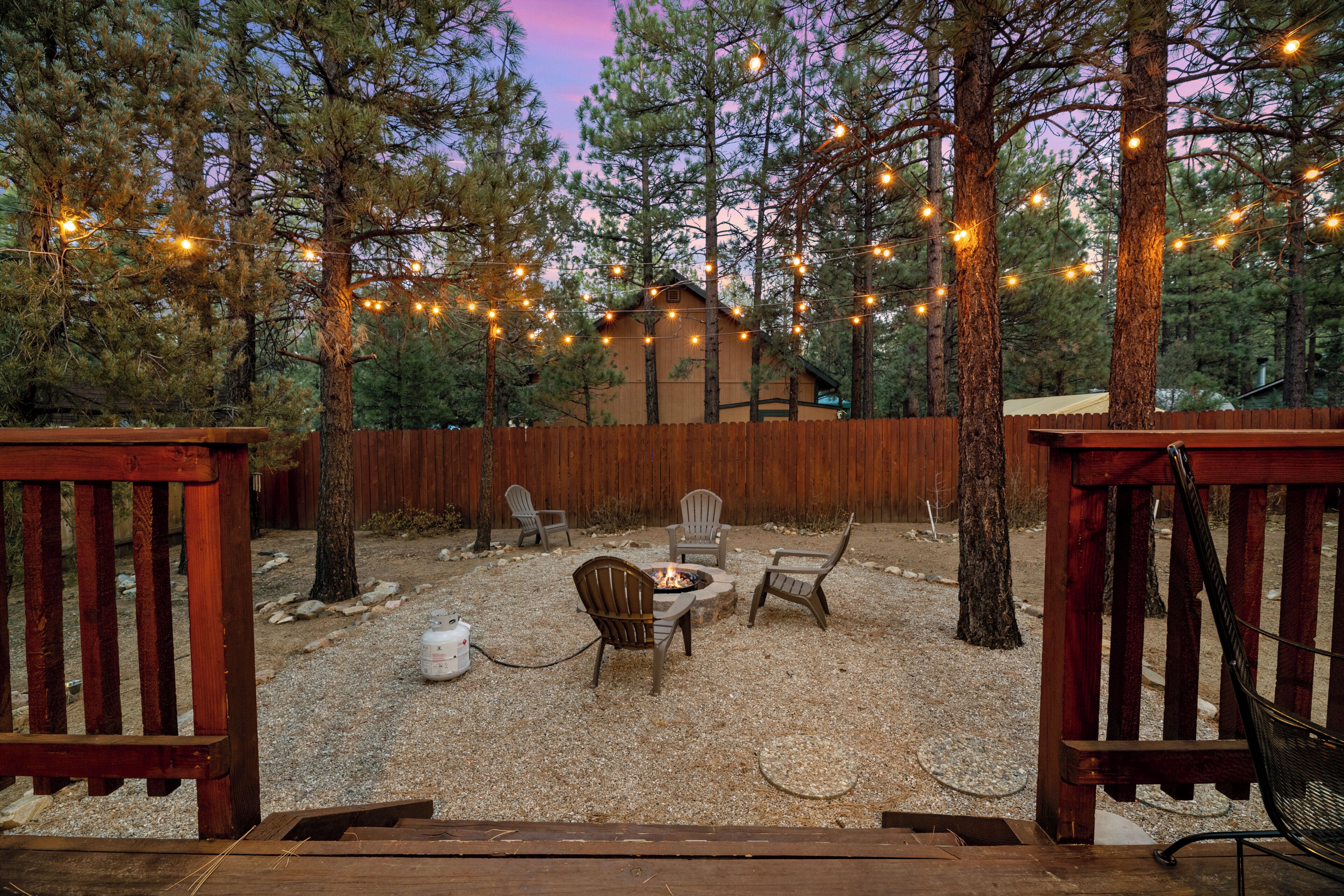 A wooden deck with a fire pit and chairs, surrounded by trees and string lights.
