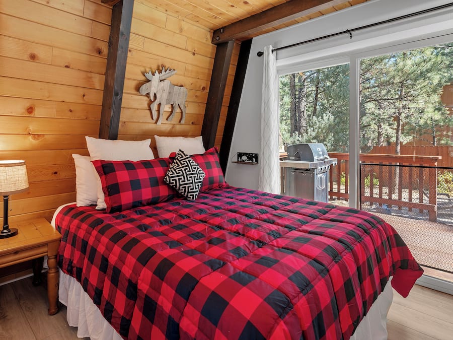A bedroom with a large bed, a wooden headboard, a nightstand with a lamp, and a view of the outdoors through a sliding glass door.