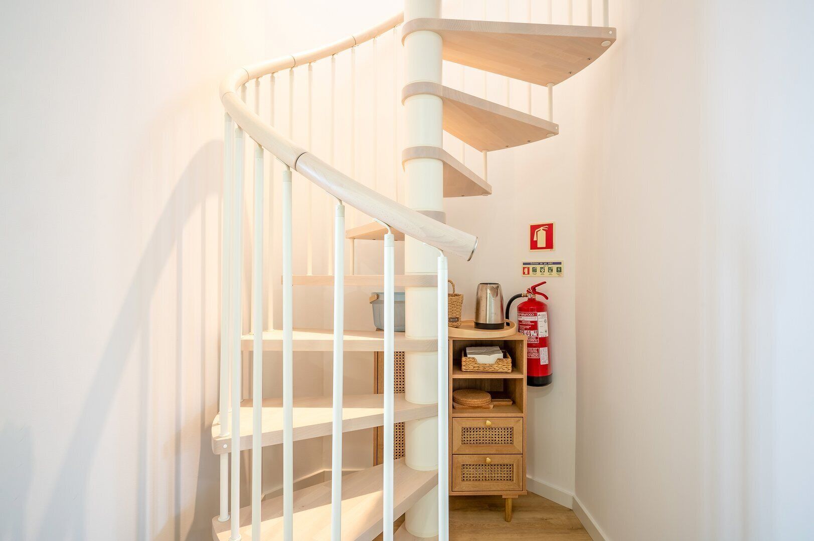 Un escalier en colimaçon blanc, avec des marches en bois et une rampe blanche. Un extincteur est fixé au mur, sur la droite.