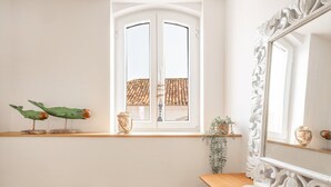A bright room with a large window, a wooden shelf with decorative items, a mirror, and a small table with a glass of milk. - GuestReady - Beachfront dream in Sesimbra (Sesimbra)