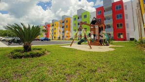 Un parque infantil con columpios y toboganes, rodeado de césped y una calle con coches estacionados.