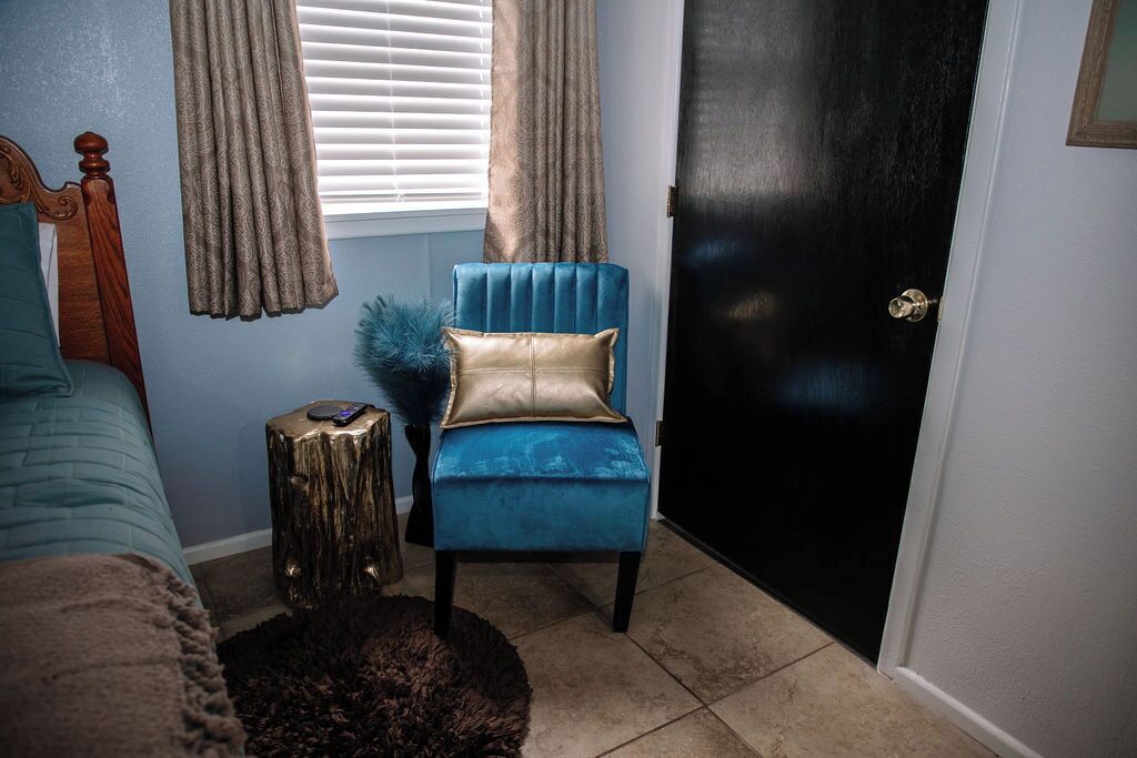 Une chambre à coucher avec un fauteuil bleu recouvert de tissu, une tête de lit en bois, une petite table ronde et une porte noire.