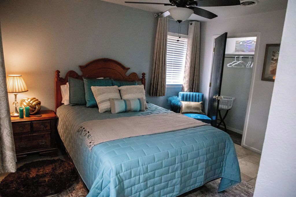 A bedroom with a bed, a nightstand, a lamp, a ceiling fan, a window with curtains, and a door with a coat rack.