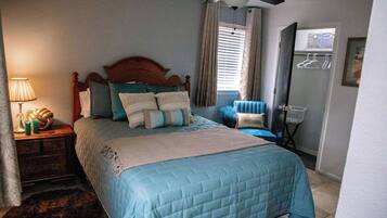A bedroom with a bed, a nightstand, a lamp, a ceiling fan, a window with curtains, and a door with a coat rack.