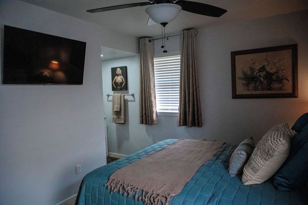 A bedroom with a bed, a ceiling fan, a window with curtains, a painting on the wall, and a framed picture above the bed.