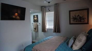 A bedroom with a bed, a ceiling fan, a window with curtains, a painting on the wall, and a framed picture above the bed.