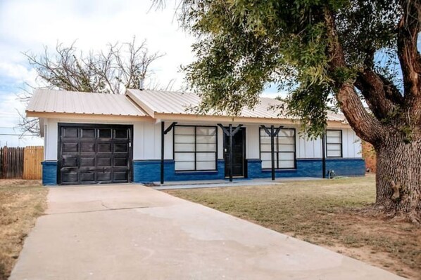 A single-story house with a garage, a front door, and a large tree in the yard. - LET D'S COZY COTTAGE #2 PROVIDE YOU A COMFORTABLE STAY IN BIG SPRING, TEXAS!!! (Big Spring)
