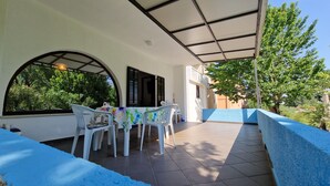 Une terrasse couverte avec une table et des chaises, un sol carrelé et une vue sur la verdure et les bâtiments.