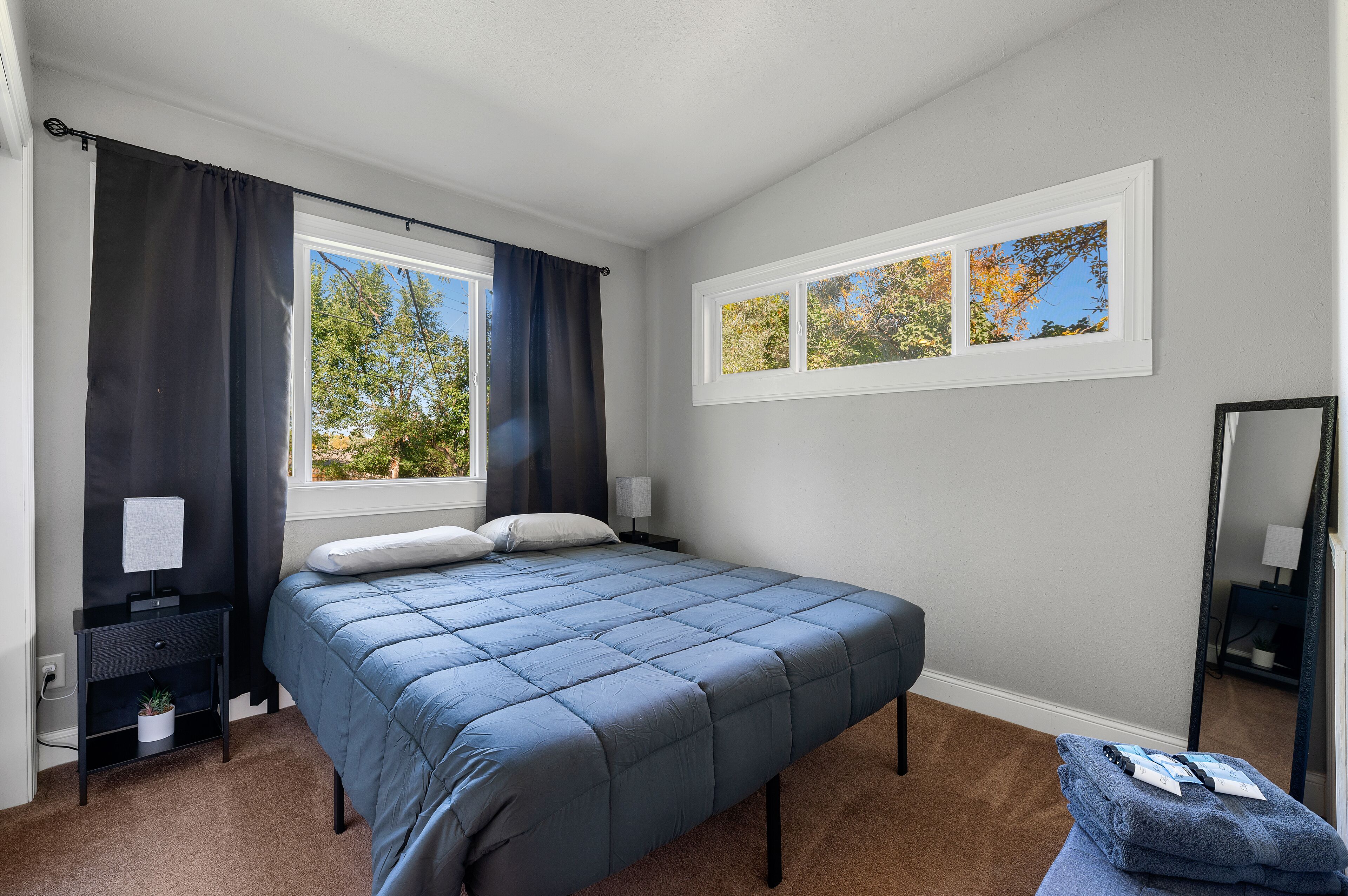 A bedroom with a bed, a nightstand, a lamp, a window with curtains, and a mirror.