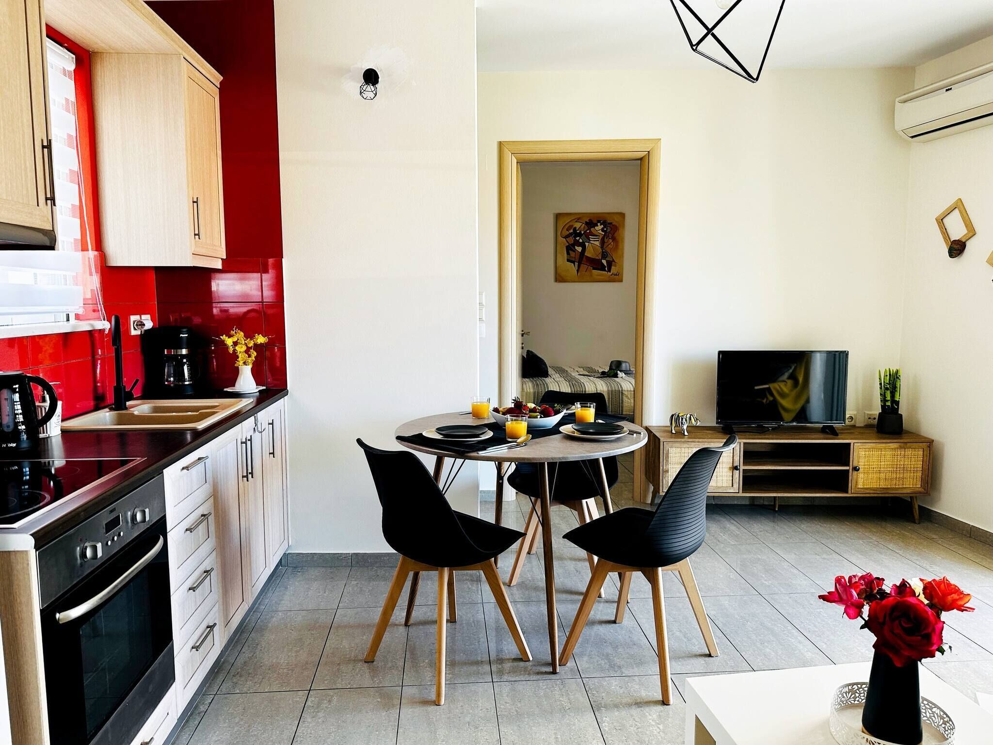 Eine moderne Küche mit roter Spritzschutz, ein Essbereich mit zwei schwarzen Stühlen, ein Fernseher auf einem Holzboden und eine kleine Pflanze auf dem Beistelltisch.
