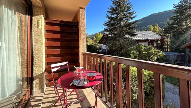 A balcony with a table and chairs, a wooden railing, and a view of a mountain and forest.